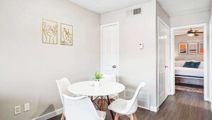 Un coin repas avec une table ronde, des chaises blanches et une plante en pot. Deux tableaux encadrés ornent le mur, et une porte blanche donne accès à une autre pièce.