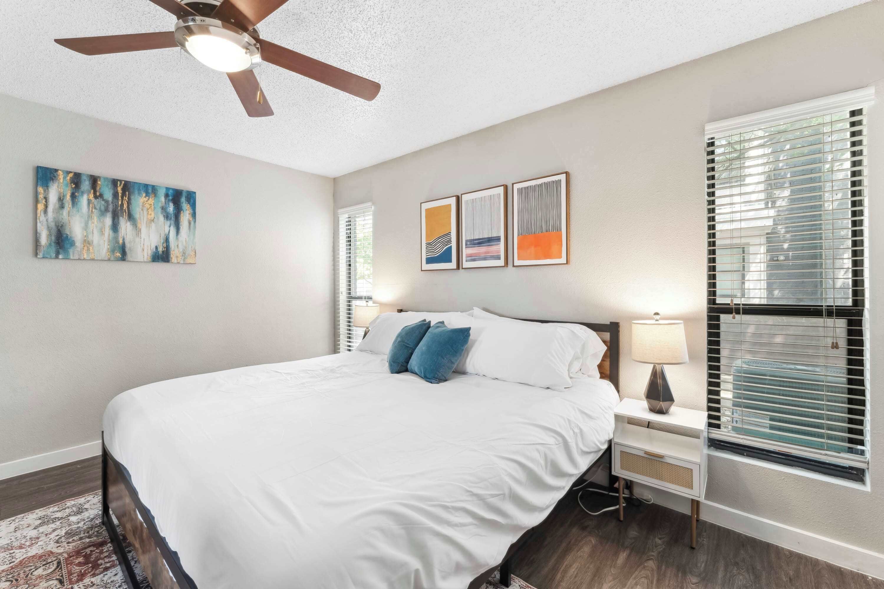 A bedroom with a bed, a ceiling fan, a nightstand with a lamp, and a window with blinds.