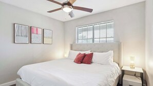 Une chambre à coucher avec un grand lit, un ventilateur de plafond, une table de chevet avec une lampe, et trois œuvres d’art encadrées accrochées au mur.