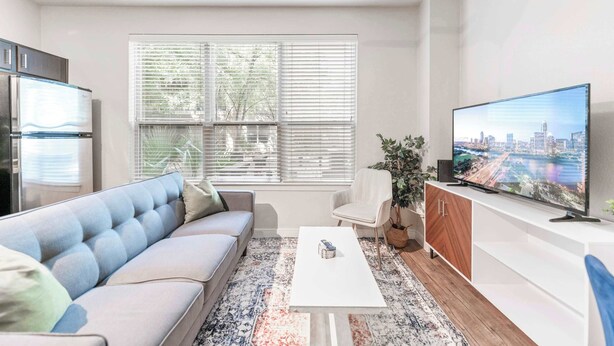 Un salón moderno con sofá, mesa de centro, televisor y un amplio ventanal con persianas.