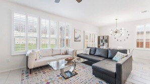 A spacious living room with two sofas, a glass coffee table, and large windows with blinds.