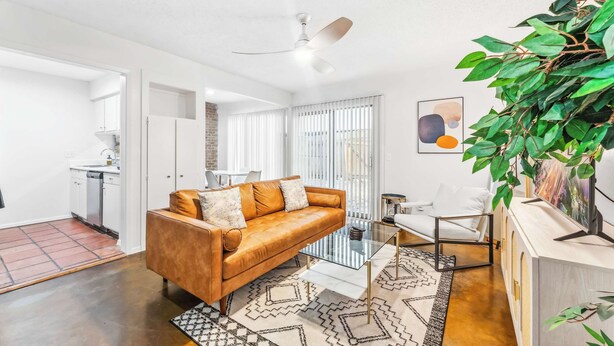 A modern living room with a brown leather sofa, a glass coffee table, and a white armchair. There is a ceiling fan, a large green plant, and a painting on the wall. - Prime SoCo cozy 1BR king suite w/ pool, parking (Austin)