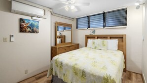 Ein Schlafzimmer mit einem Bett, einem Deckenventilator, einem Fenster mit Jalousien, einem Nachttisch, einem Spiegel und einer Wand-Klimaanlage.