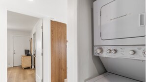 A laundry room with a white Whirlpool washer and dryer, a wooden door, and a small wooden cabinet with a television.