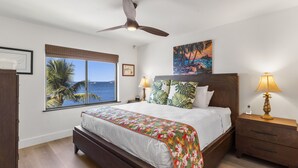 A bedroom with a bed, a ceiling fan, a window with a view of the sea, and a painting on the wall. - Kihei Beach, #510 2 Bedroom Condo by RedAwning (Kihei)