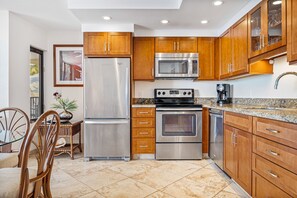 A kitchen with stainless steel appliances, wooden cabinets, a granite countertop, and a dining area with wicker chairs. - Kihei Beach, #406^ 1 Bedroom Condo by RedAwning (Kihei)
