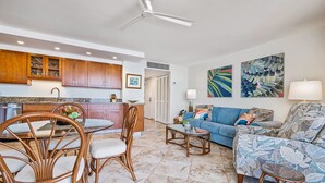 A modern kitchen with wooden cabinets, a dining area with wicker chairs, a living room with a sofa and patterned cushions, and a ceiling fan. - Kihei Beach, #406^ 1 Bedroom Condo by RedAwning (Kihei)
