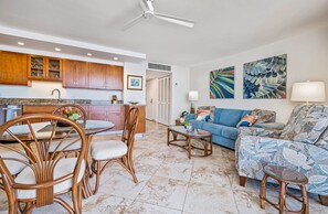 A modern kitchen with wooden cabinets, a dining area with wicker chairs, a living room with a sofa and patterned cushions, and a ceiling fan. - Kihei Beach, #406^ 1 Bedroom Condo by RedAwning (Kihei)
