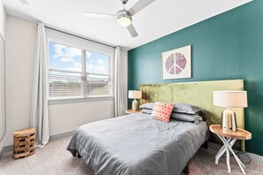 Una camera da letto con un letto, due comodini con lampade, una ventola a soffitto, una finestra con tende e una parete verde decorativa.