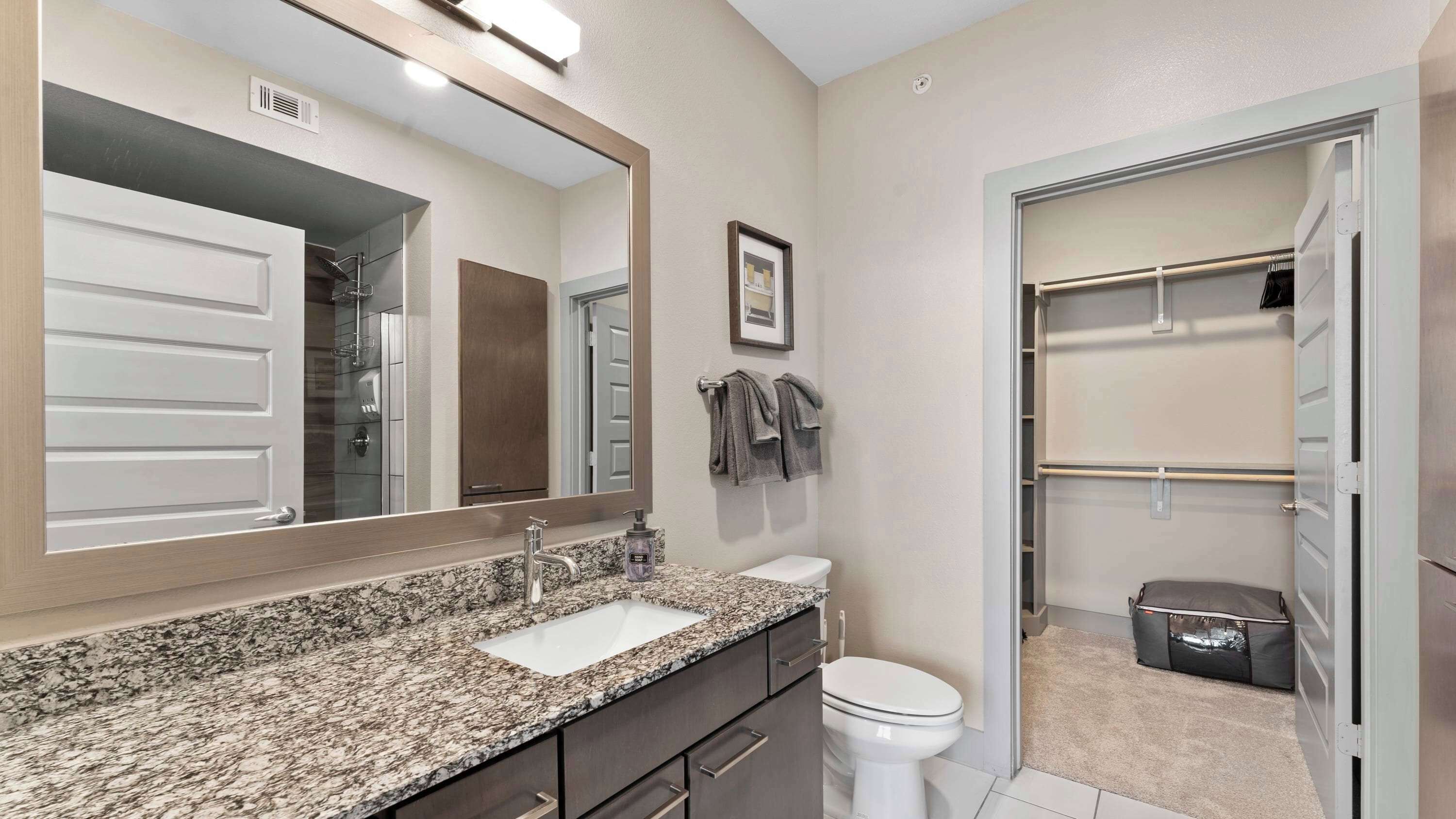 A bathroom with a granite countertop sink, a toilet, a large mirror, and a walk-in closet with hanging clothes.
