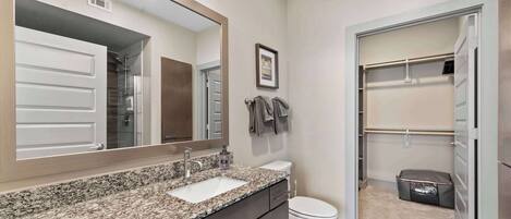 A bathroom with a granite countertop sink, a toilet, a large mirror, and a walk-in closet with hanging clothes.