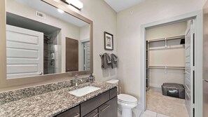 Un bagno moderno con lavabo su piano in granito, uno specchio grande, un WC e una cabina armadio con abiti appesi.