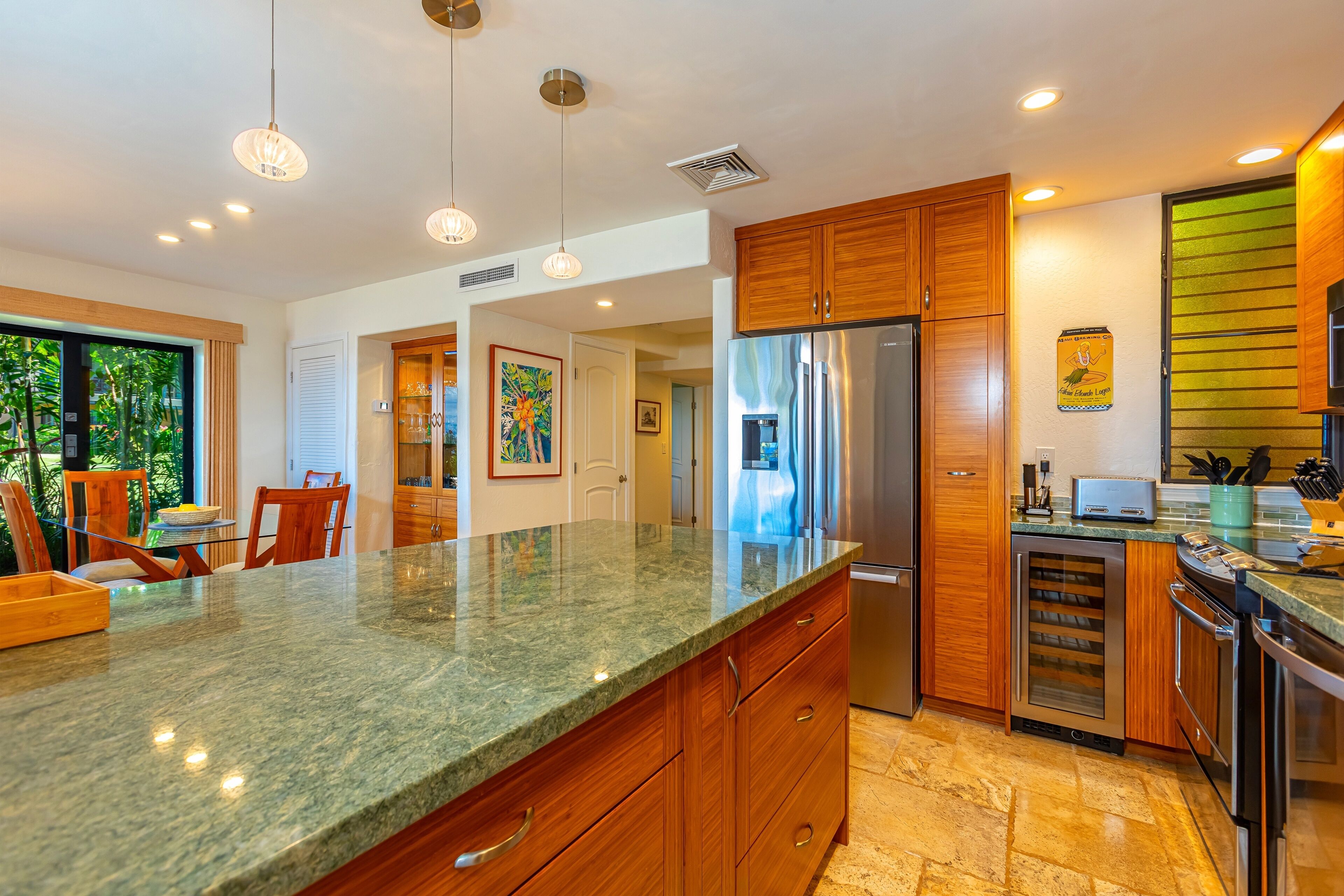 A modern kitchen with granite countertops, wooden cabinets, stainless steel appliances, pendant lighting, and a view of greenery outside.
