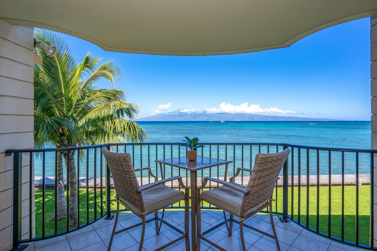 Un balcon avec deux fauteuils en osier et une petite table, d’où l’on peut admirer une mer d’un bleu profond et une chaîne de montagnes lointaine.