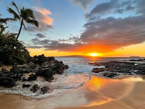 A beach with rocky terrain, palm trees, and a sunset.
