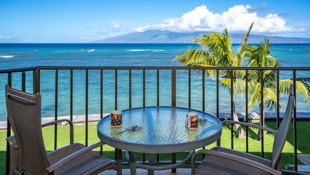 Ein Balkon mit einem Glastisch, zwei Stühlen und Blick auf das Meer und ein Gebirge.