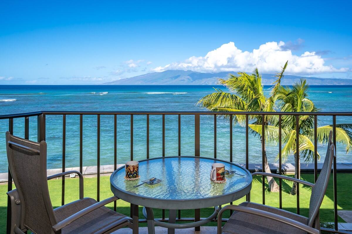 Un balcon avec une table en verre, deux chaises et une vue sur l’océan et une montagne.