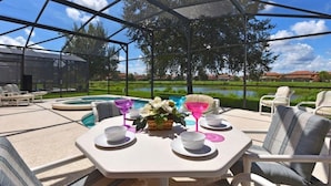 An outdoor patio with a table set for dining, surrounded by lounge chairs, a pool, and a view of a landscaped area. - Villa Marcello 4 Bedroom Home by RedAwning (Kissimmee)
