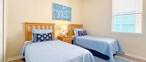 A bedroom with two beds, a ceiling fan, a window with blinds, and a wall-mounted artwork.