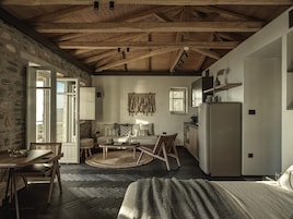 A rustic room with exposed wooden beams, a stone wall, a sofa, a dining table, and a kitchen area.