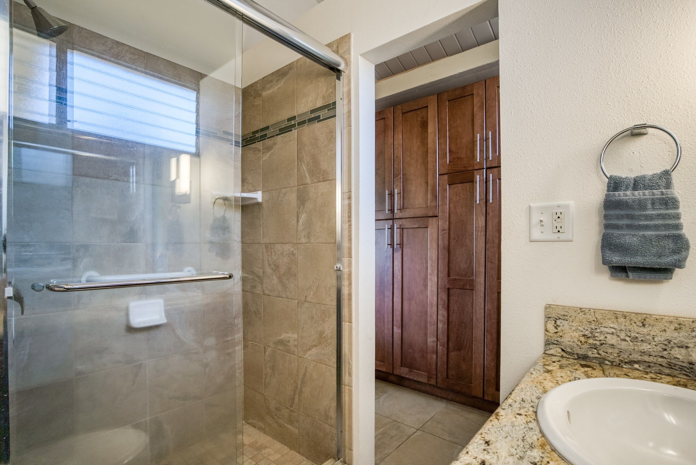 A bathroom with a glass shower enclosure, a granite countertop, a sink, wooden cabinets, and a towel ring.