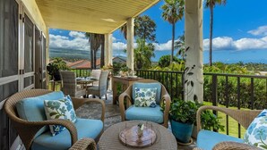 A covered outdoor patio with wicker furniture, a round table, and a view of palm trees and distant hills. - Maui Hill by Coldwell Banker Island Vacations (Kihei)