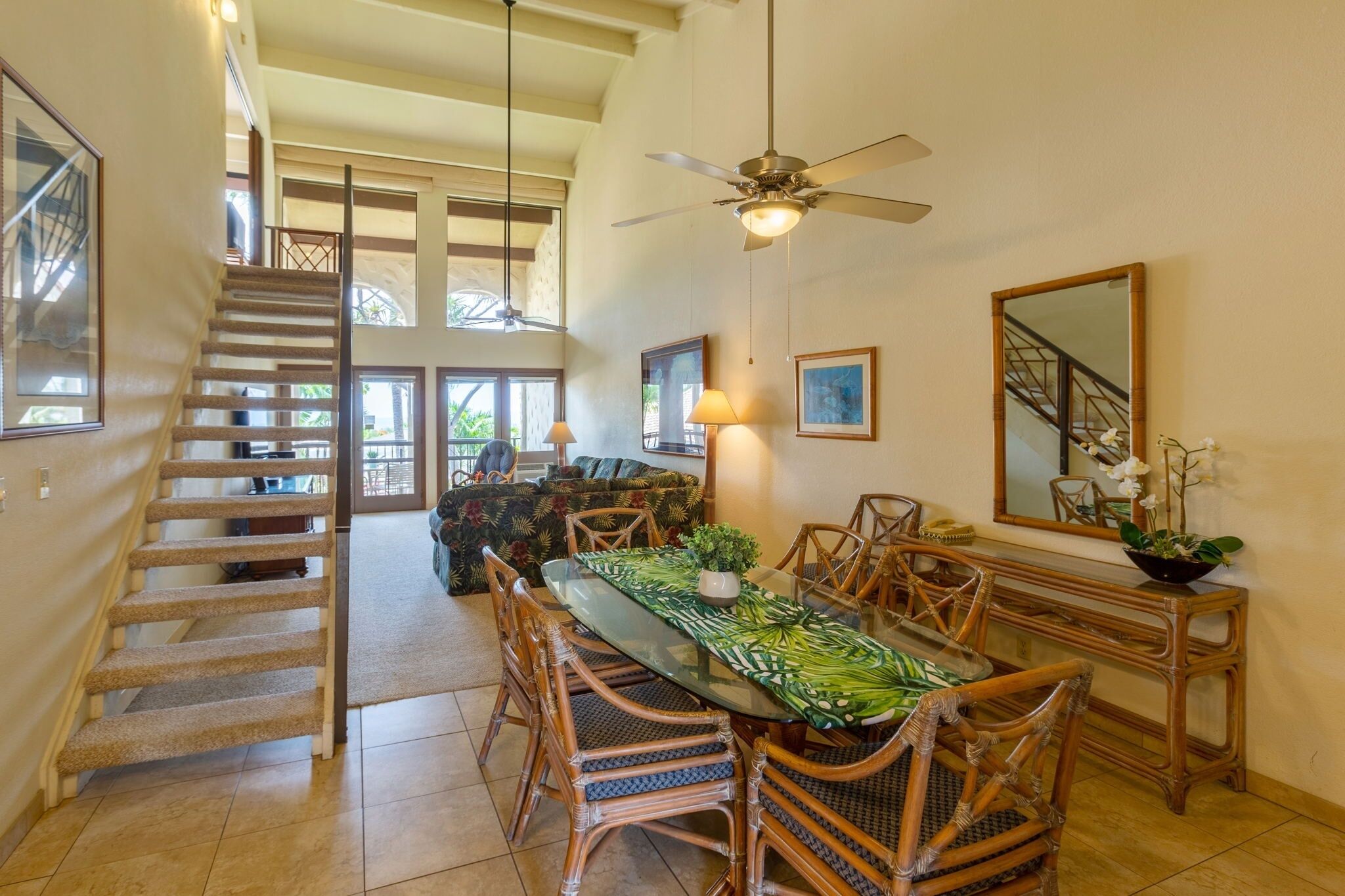 A spacious dining area with a glass-top table, wicker chairs, and a ceiling fan. A staircase leads to an upper level with large windows.