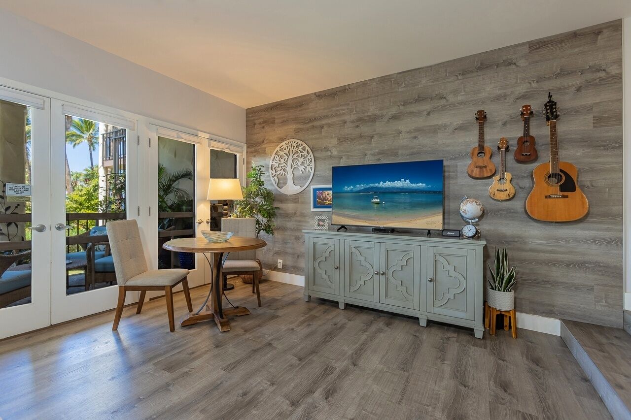 Un salon avec un plancher en bois, une table ronde entourée de chaises, un téléviseur, un meuble et une collection de guitares accrochées au mur.