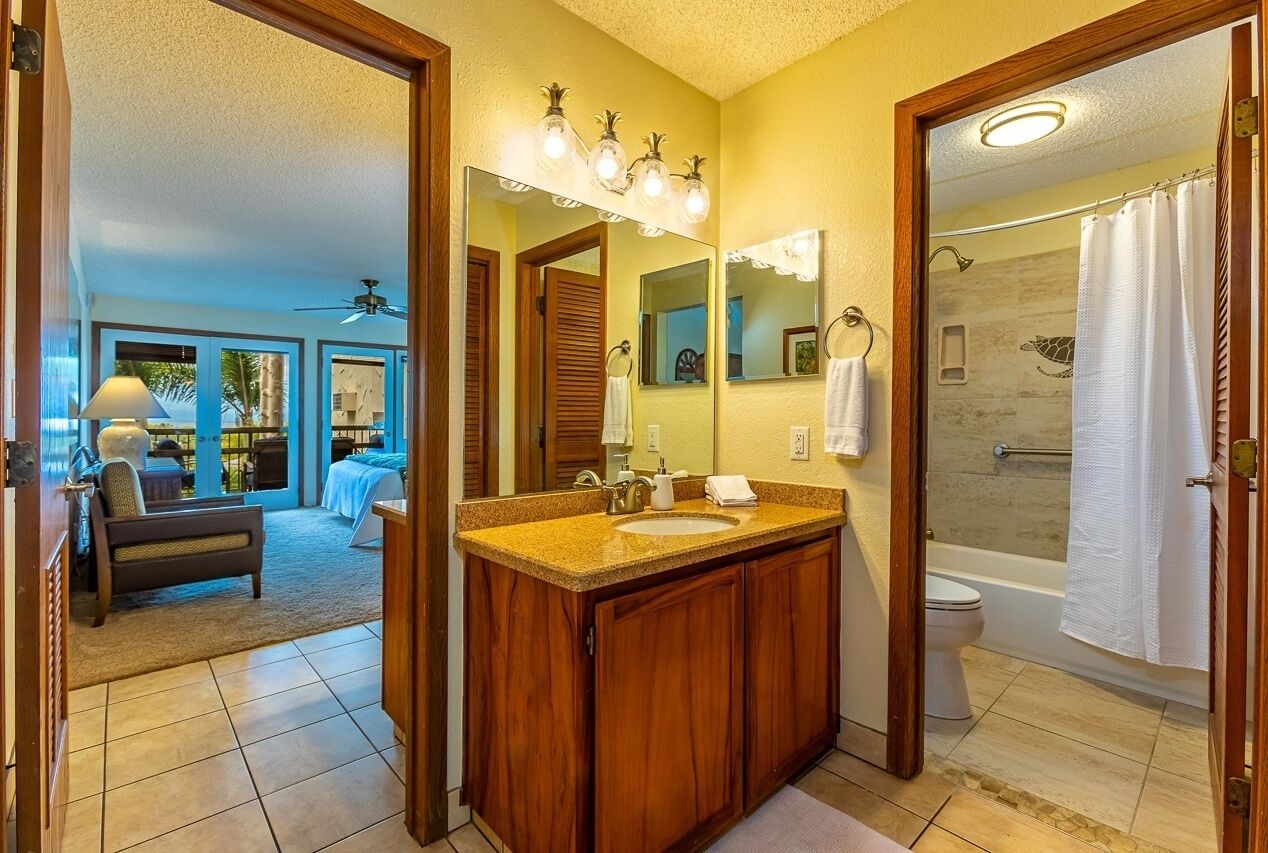 A bathroom with a wooden vanity, a large mirror, a shower, and a view of a living area with a sofa and a ceiling fan.
