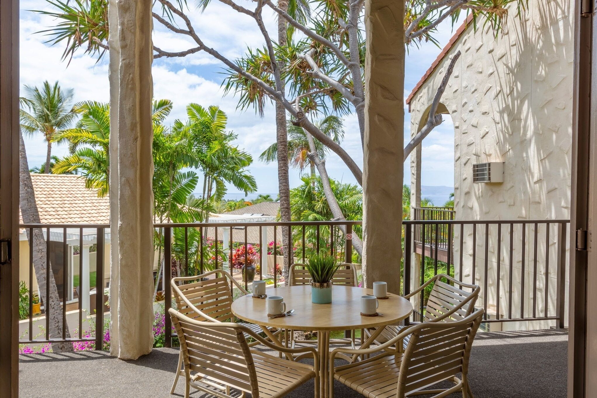 A round table with six chairs and a potted plant in the center, surrounded by a balcony with a view of palm trees and a building.