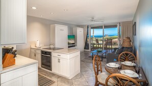 A modern kitchen with white cabinetry, a dining area with wooden chairs, and a view of the ocean through large sliding doors.