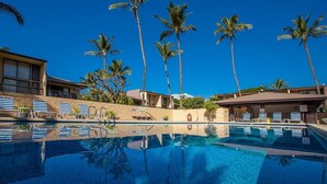 Zona de piscina con sillones de descanso, palmeras y un edificio de fachada de piedra.
