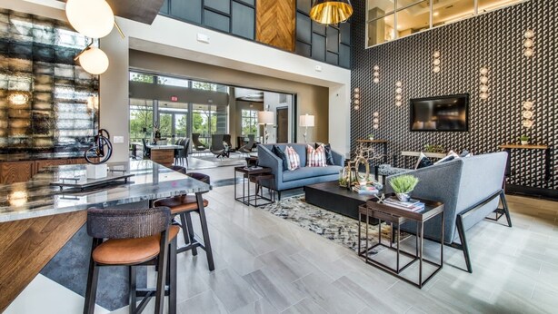 Een ruime lobby met een hoge plafond, een centraal zithoek met banken en stoelen, een bar met krukken, en een grote televisie aan de muur.