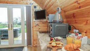 Interior acogedor de una cabaña, con paredes de madera, una zona de cocina con estufa y televisor, y una mesa para el desayuno con croissants, uvas y jugo de naranja.