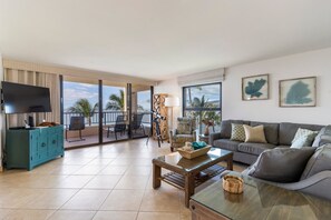 Een woonkamer met een bank, een salontafel en een televisie. Er is een balkon met stoelen en uitzicht op palmbomen.