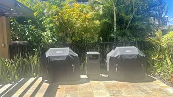 Two Weber barbecue grills on a tiled patio surrounded by lush greenery and a metal fence.