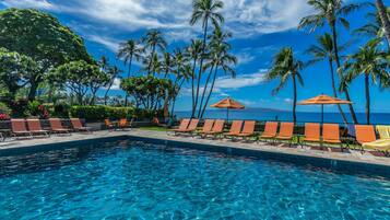 A pool with clear blue water, surrounded by lounge chairs and palm trees, with a view of the ocean.
