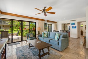 A spacious living room with a ceiling fan, a sofa, a coffee table, and a view of a tropical garden through large sliding glass doors.