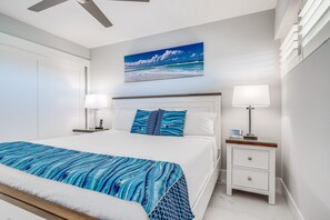 A bedroom with a bed, bedside tables, a nightstand, a painting of a beach scene, and a ceiling fan. - Sugar Beach Resort, #ph27 1 Bedroom Condo by RedAwning (Kihei)