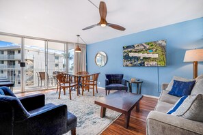 A modern living room with a dining area, a ceiling fan, and a large window overlooking a building. - Kamaole Nalu, #601 2 Bedroom Condo by RedAwning (Kihei)