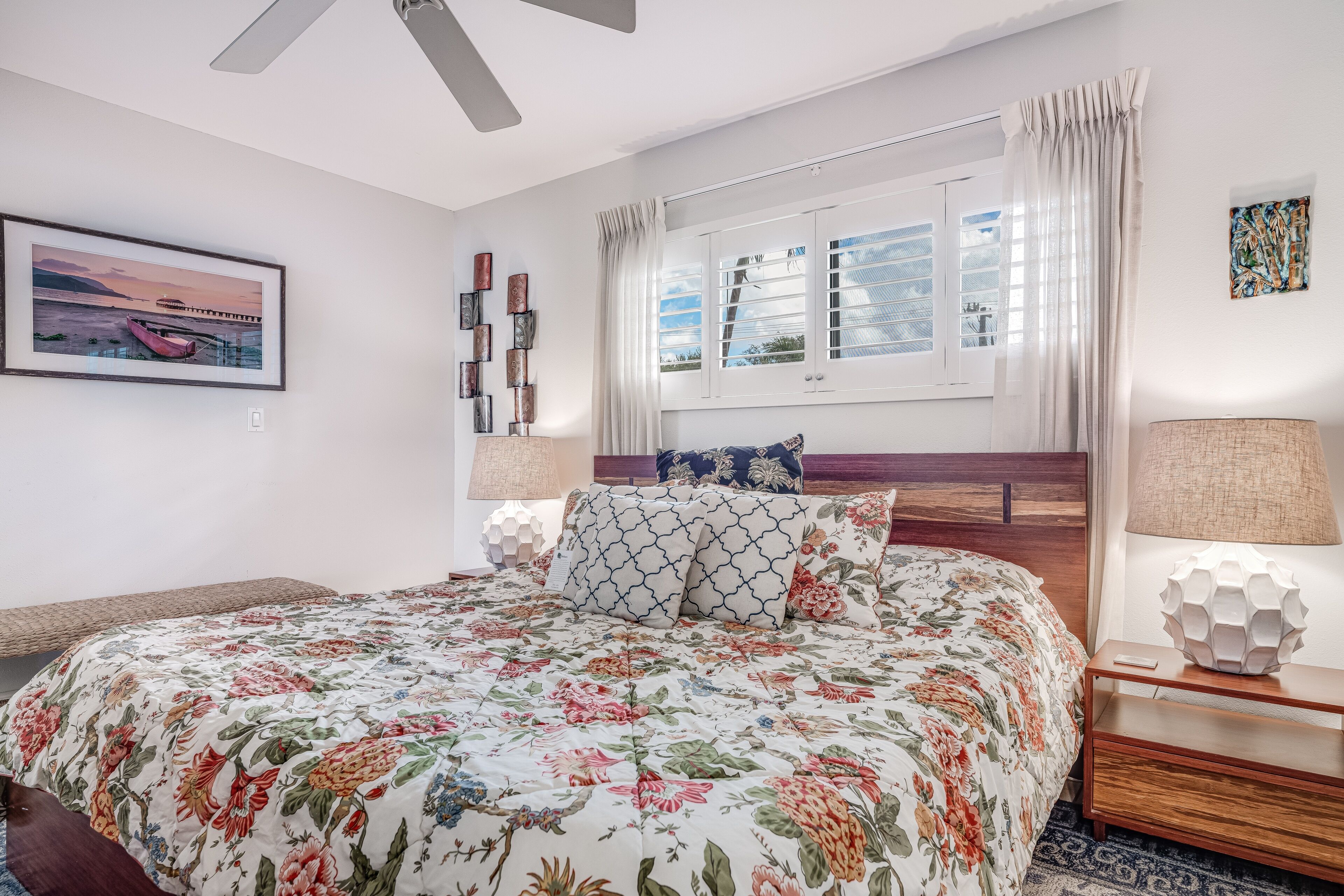 A bedroom with a bed, a window with curtains, a wall-mounted picture, and a bedside table with a lamp.