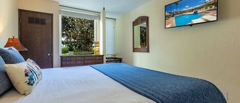 A bedroom with a bed, a ceiling fan, a TV, a window with blinds, and a view of a pool outside.