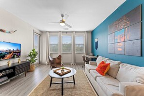A modern living room with a flat-screen TV, a ceiling fan, a sofa, a coffee table, and a large window with blinds.