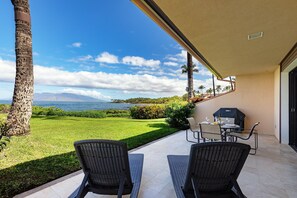 A patio with two wicker chairs, a table, and a grill, overlooking a grassy area with palm trees and a view of the ocean. - Makena Surf, #f-110 2 Bedroom Condo by RedAwning (Kihei)