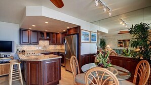A modern kitchen with a central island, wooden cabinets, stainless steel appliances, and a dining area with wicker chairs. - Kihei Beach, #307 1 Bedroom Condo by RedAwning (Kihei)