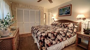 A bedroom with a bed, a ceiling fan, a nightstand with a lamp, a dresser, a mirror, and a door with a view of the outside. - Kihei Beach, #307 1 Bedroom Condo by RedAwning (Kihei)