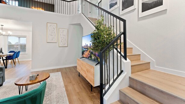 A spacious living area with a staircase, wooden flooring, a dining area with blue chairs, and a TV on a wooden console.