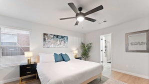 A bedroom with a bed, bedside tables, a ceiling fan, a window with blinds, and a door.