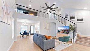 A spacious living area with a grey sofa, a dining table with chairs, a staircase, and a cityscape TV.