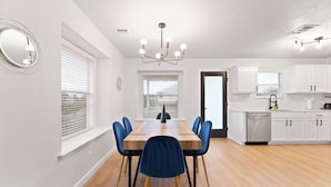 A modern kitchen with white cabinets, a stainless steel dishwasher, and a wooden dining table with blue chairs.